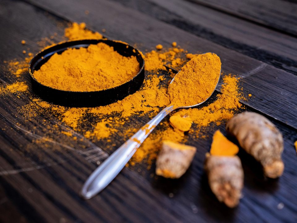 Turmeric powder in a black dish with a spoon and dried rhizomes on dark wood